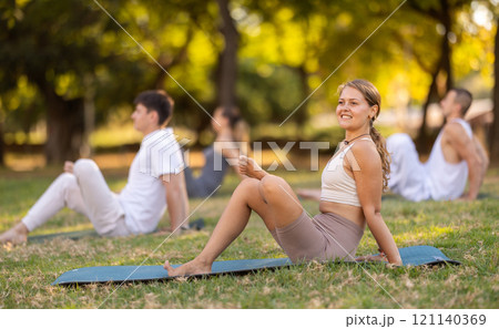 Yoga lesson visitors with girl coach perform halasana in park 121140369