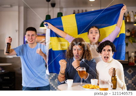 Group of happy friends, sports supporters cheering for favorite team with flag of Sweden while watching match on TV in pub Group of happy friends, sports supporters cheering for favorite team with flag of Sweden while watching match on TV in pub 121140575