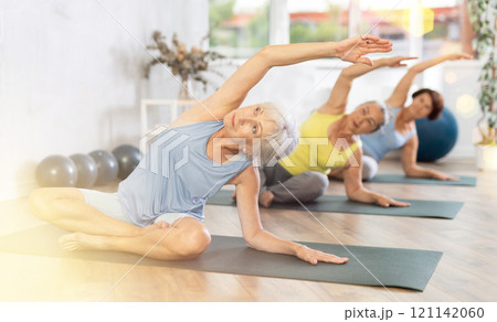 Old woman practicing lotus pose of yoga with side bend in training room 121142060