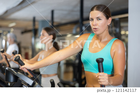 Slender athletic girl during cardio exercises on elliptical simulator in fitness center. 121142117