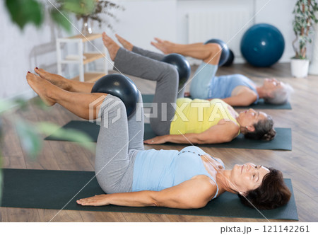 Old woman practicing pilates with softball between legs in light fitness room Old woman practicing pilates with softball between legs in light fitness room 121142261