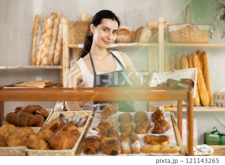 Young woman employee puts croissants in window, arranges display of goods at bakery. 121143265