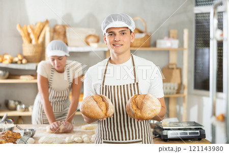 Young man are standing in kitchen with finished products two loaves of rye bread Young man are standing in kitchen with finished products two loaves of rye bread 121143980