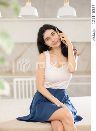 Woman talking on smartphone while sitting on kitchen table Woman talking on smartphone while sitting on kitchen table 121146587