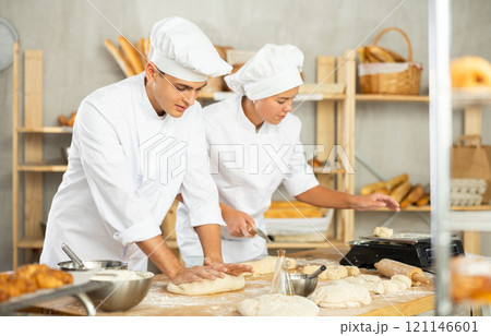 Two young bakers prepare dough in kitchen of bakery - together they knead dough, cut it with knife and weigh it on scales Two young bakers prepare dough in kitchen of bakery - together they knead dough, cut it with knife and weigh it on scales 121146601