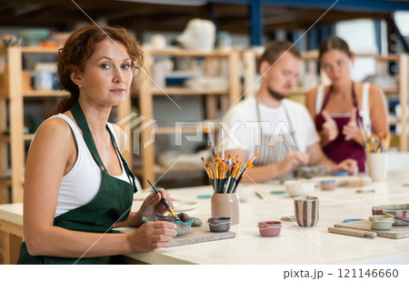 Woman learns to make designs on clay plate using brush and paint in pottery workshop Woman learns to make designs on clay plate using brush and paint in pottery workshop 121146660