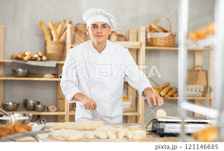 Young male baker cutting and weighing dough on scales in bakehouse 121146685