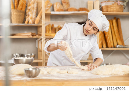 Young woman works as baker, mold baguette from dough, creates long loafs from dough. Young woman works as baker, mold baguette from dough, creates long loafs from dough. 121147001