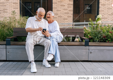 Portrait of cheerful elderly couple laughing sitting on city bench, sharing funny moment on smartphone, embodying senior love and togetherness through modern technology. Concept of retirement life Portrait of cheerful elderly couple laughing sitting on city bench, sharing funny moment on smartphone, embodying senior love and togetherness through modern technology. Concept of retirement life 121148309