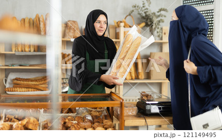 Selling fresh bread in an oriental bakery - girl seller hands bag of baguettes to female customer 121149495