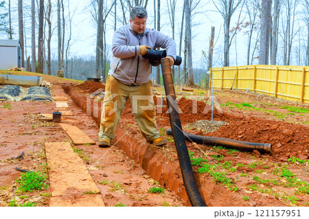 Man is working on installing drainage system in backyard, in freshly dug trench to enhance water flow management. 121157951