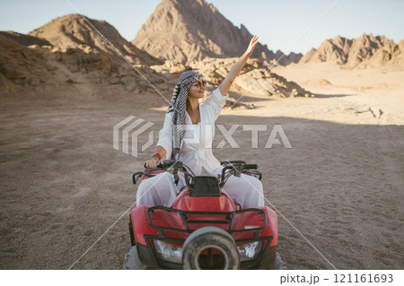 A rather attractive woman is going on a safari, driving an ATV in the desert. Beautiful young woman with a scarf on her head sits on a red four-wheeled ATV and rides towards adventure 121161693