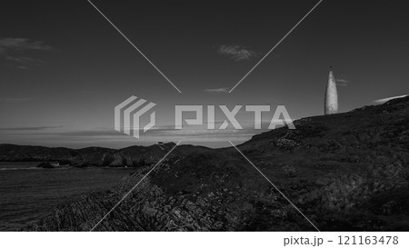 A white, tall, slender marker stands on a grassy cliff. Dark, rugged rocks form the cliff face. Sea stretches to a distant coastline. A white, tall, slender marker stands on a grassy cliff. Dark, rugged rocks form the cliff face. Sea stretches to a distant coastline. 121163478