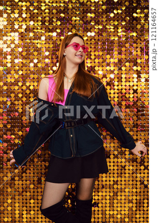 Young woman posing confidently in trendy outfit against a sparkling golden backdrop 121164657