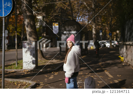 A young woman strolls through colorful autumn streets in a casual outfit and pink beanie 121164711