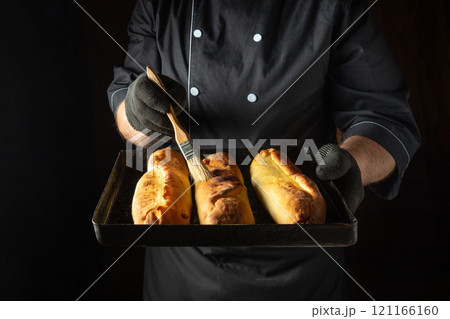 A skilled baker holds a tray of freshly baked loaves, gently brushing them with melted butter. The warm glow of the kitchen enhances the inviting atmosphere during the evening 121166160