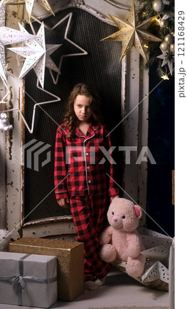 Portrait of sad dissatisfied little girl in night pajamas holding teddy bear in hands. Child disappointed with gifts from Santa Claus. Christmas night, festive atmosphere 121168429