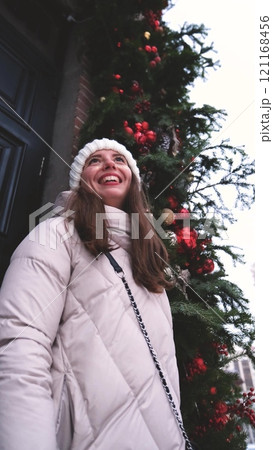 Portrait of a beautiful young woman in winter clothes. The woman smiles happily against the background of a snowy street traditionally decorated for Christmas holidays Portrait of a beautiful young woman in winter clothes. The woman smiles happily against the background of a snowy street traditionally decorated for Christmas holidays 121168456
