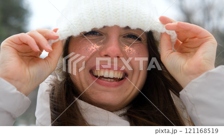 Woman in warm white knitted hat pulled down over eyes, smiling and enjoying snow, laughing and looking at camera. Active winter recreation. Wide, beautiful smile. Close-up 121168519