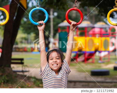 Happy girl hanging on monkey bar by hand doing exercise. Little Asian girl playing at outdoor playground in the park on summer vacation. Healthy activity. Happy girl hanging on monkey bar by hand doing exercise. Little Asian girl playing at outdoor playground in the park on summer vacation. Healthy activity. 121171992