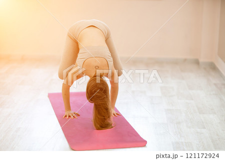 A young woman practices yoga in the gym. A girl with long hair and a beige tracksuit stands in a crow pose on a pink rug. A woman performs bokasana. A young woman practices yoga in the gym. A girl with long hair and a beige tracksuit stands in a crow pose on a pink rug. A woman performs bokasana. 121172924