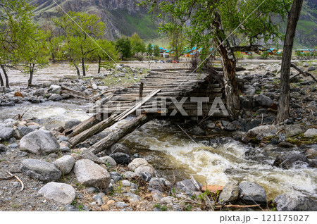An old, flimsy wooden bridge across a turbulent, full-flowing mountain river with a fast current. 121175027