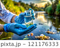 Hands in protective gloves hold a flask with water samples on a blurred background of a pond. 121176333