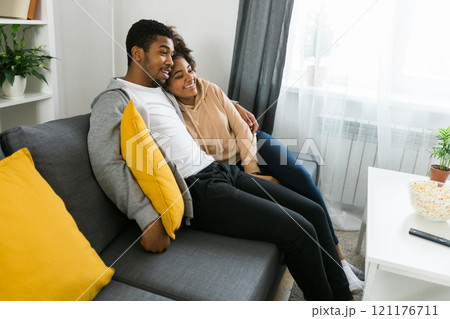 African American couple relaxes on a cozy sofa, sharing a joyful moment while watching a show. Essence of quality time and comfort in a stylish modern living room 121176711
