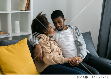 African American couple sits on a cozy gray sofa, embracing each other and enjoying a peaceful moment at home. Love and intimacy and a sense of comfort in a modern and inviting interior 121176765