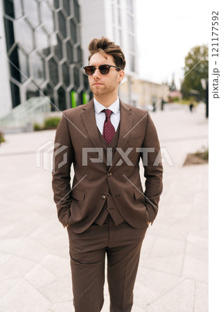 Vertical portrait of confident young businessman in stylish suit walking in downtown, exuding success and professionalism. Determined ambitious business man in sunglasses embodying modern leadership. 121177592
