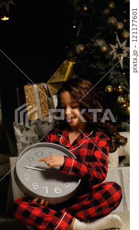 Little girl in night pajamas holding a watch in her hands waiting for Santa Claus and magic. Christmas night, festive atmosphere. 121177601