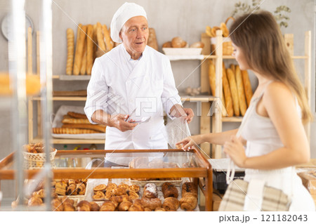 Beautiful interested young girl buyer points to products on display and chooses baked goods, elderly man seller helping her in bakery store 121182043