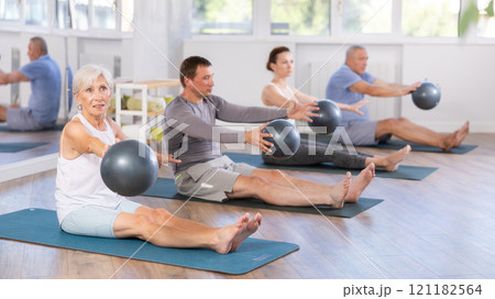 Group of mature people in sportswear exercising Pilates with soft balls while sitting on mats in fitness studio 121182564