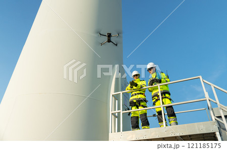 Two wind turbine or windmill workers or engineer stand on base of the pole and one control drone with discuss together in workplace field. Two wind turbine or windmill workers or engineer stand on base of the pole and one control drone with discuss together in workplace field. 121185175