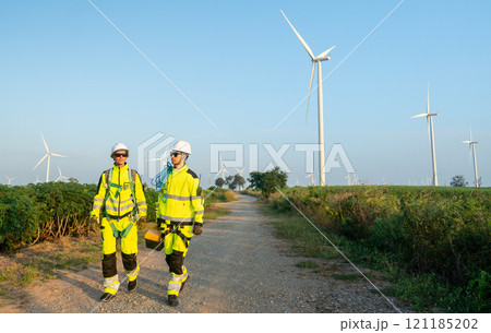 Wide shot of two of wind turbine or windmill workers or technicians carry tools and walk along the road in workplace field with row of wind turbine in the background. Wide shot of two of wind turbine or windmill workers or technicians carry tools and walk along the road in workplace field with row of wind turbine in the background. 121185202