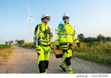 Two of wind turbine or windmill workers or technicians carry tools and walk along the road in workplace field with row of wind turbine in the background. Two of wind turbine or windmill workers or technicians carry tools and walk along the road in workplace field with row of wind turbine in the background. 121185204