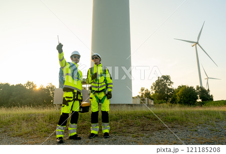 Two of wind turbine or windmill workers or technician stand on base of wind turbine and one point to their right side with warm light from sunset in the background. 121185208