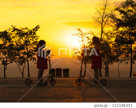 Silhouette of cute sisters riding scooters in the park during a summer sunset at a public park. Active leisure and outdoor sport for child. Silhouette of cute sisters riding scooters in the park during a summer sunset at a public park. Active leisure and outdoor sport for child. 121186355