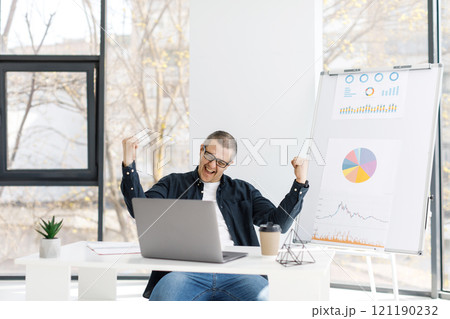 A man dressed in casual clothes works in the office with a laptop and expresses positive emotions 121190232