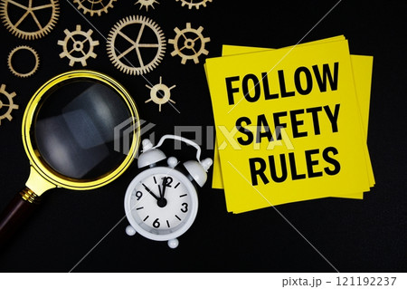 Follow Safety Rules text message on sticky note with magnifying glass and alarm clock on black background 121192237
