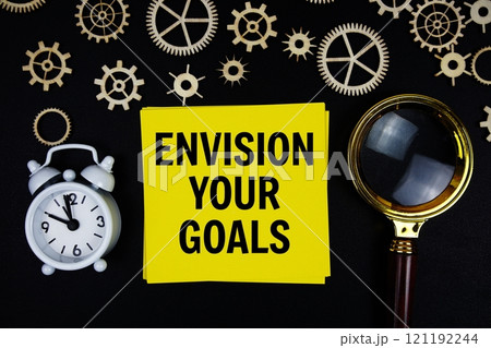 Envision Your Goals text message on sticky note with magnifying glass and alarm clock on black background 121192244