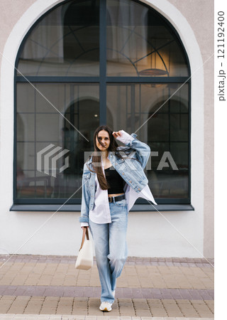 Young woman in a black top and blue jeans is walking down a brick sidewalk. She is carrying a white purse and has her hand up in the air. Concept of casual elegance and confidence 121192400