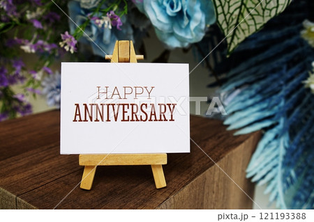 Happy Anniversary text write on paper card with wooden easel and flowers decoration on wooden background 121193388