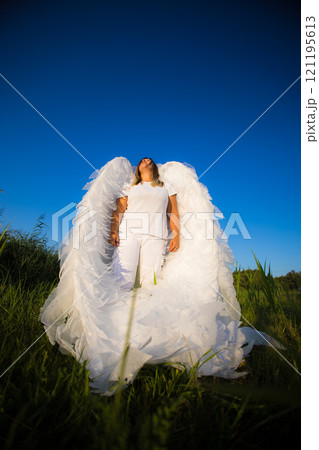 A woman with large white angel wings stands on a green field, throwing her head back and looking at the clear blue sky. The angel looks into the bottomless blue sky. 121195613