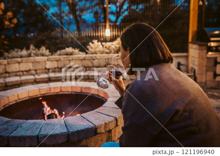 Elderly woman sitting alone by fire pit, drinking glass of red wine. Elderly woman sitting alone by fire pit, drinking glass of red wine. 121196940