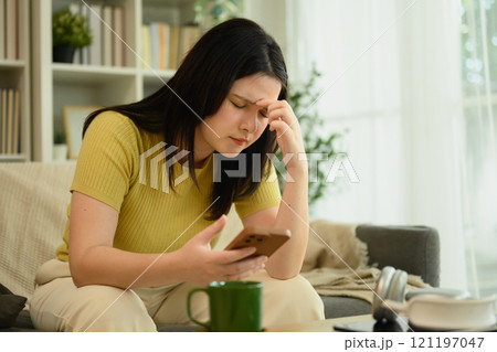 Young woman holding smartphone and touching head with closed eyes, feeling headache or exhaustion 121197047