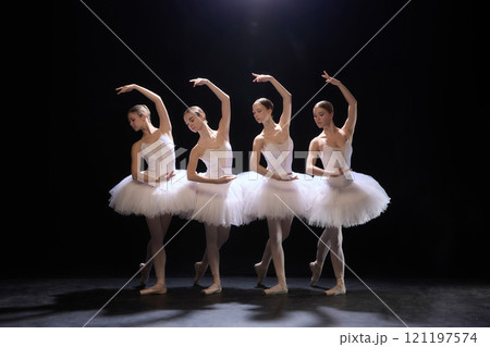 Four young and beautiful ballerinas in white tutus showing perfect symmetry during graceful performance eon dimly lit stage against black background Four young and beautiful ballerinas in white tutus showing perfect symmetry during graceful performance eon dimly lit stage against black background 121197574