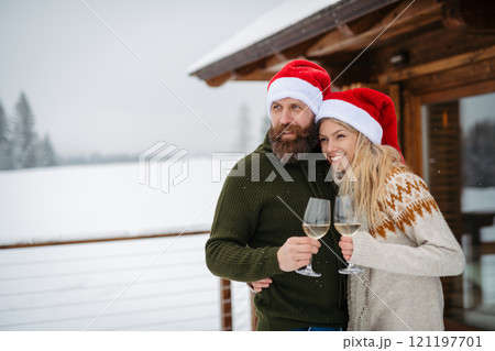 Mature couple wearing Santa hats, standing in front cabin and toasting with wine glasses Mature couple wearing Santa hats, standing in front cabin and toasting with wine glasses 121197701