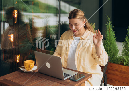 Happy 30s woman doing business on laptop and smiles. Young woman enjoy her work outdoor. Remote work, freelance. Young pretty female talking on video call and waving hand in cafe Happy 30s woman doing business on laptop and smiles. Young woman enjoy her work outdoor. Remote work, freelance. Young pretty female talking on video call and waving hand in cafe 121198323