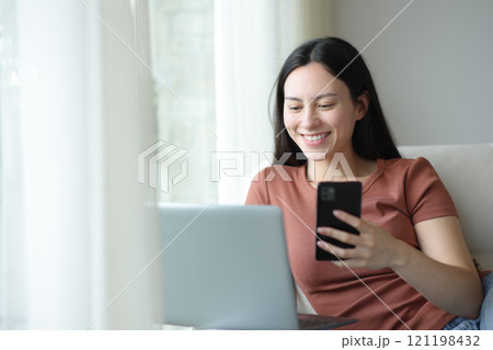 Happy asian woman using multiple devices sitting in a sofa Happy asian woman using multiple devices sitting in a sofa 121198432